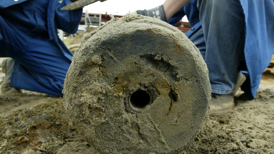 Zwei Personen in Arbeits- beziehungsweise Schutzkleidung, die eine alte Bombe ausgegraben haben.