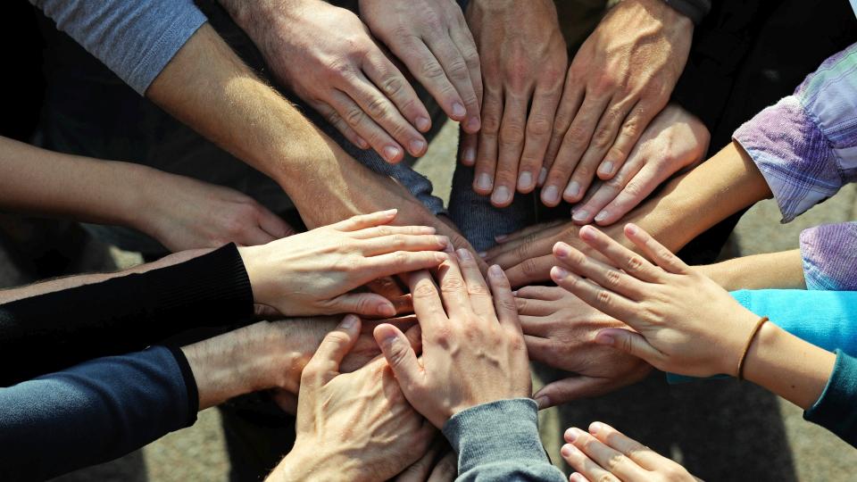 Mehrere Personen stehen zusammen in einem geschlossenen Kreis und halten jeweils einen Arm in die Mitte die Kreises, in der sich alle Hände berühren. 
