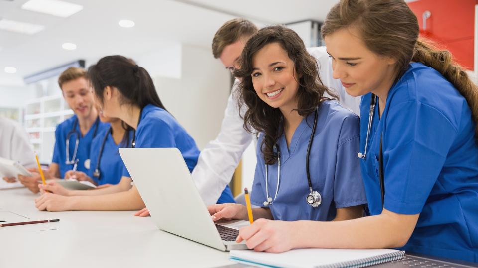 Junge Ärztinnen und Ärzte sitzen lernend an einem Tisch. Zwei Ärztinnen schauen dabei auf einen aufgeklappten Laptop. Der stehende Arzt im Hintergrund fungiert wahrscheinlich als Ansprechperson.