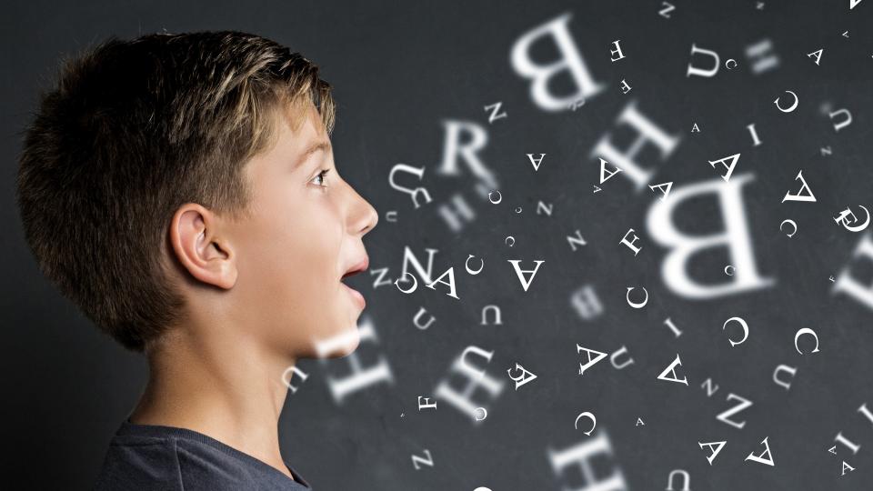Eine Junge steht seitlich vor einer Tafel, aus seinem Mund strömen Buchstaben.