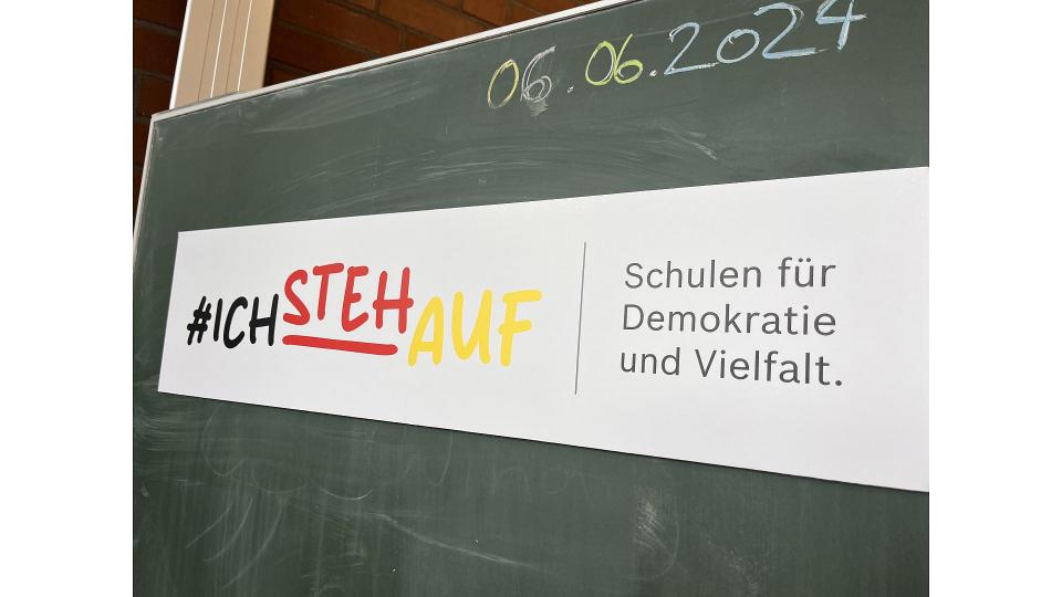 Abgebildet ist ein Banner mit der Aufschrift "#IchStehAuf", der auf eine Schultafel geklebt wurde.
