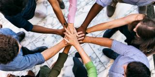 Eine Gruppe von jungen Erwachsenen, die unterschiedlicher ethnischer Herkunft sind und gemeinsam in einem geschlossenen Kreis stehen, in dessen Mitte sich die Hände der Personen berühren.