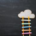 Eine Leiter aus Bleistiften und farbigen Kreidestücken zu einer Wolke vor einer Tafel.