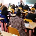 Schülerinnen und Schüler sitzen an Einzeltischen in einem Klassenraum und schreiben eine Prüfung. Vor den Schülerinnen und Schülern steht eine Aufsichtsperson. 