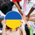 Abgebildet ist eine Frau, die eine große Sprechblase auf Kopfhöhe hält. Farblich ist die Sprechblase an die Flagge der Ukraine angelehnt. Im Hintergrund legen im Kreis stehende Personen jeweils eine Hand über die einer anderen Person. 